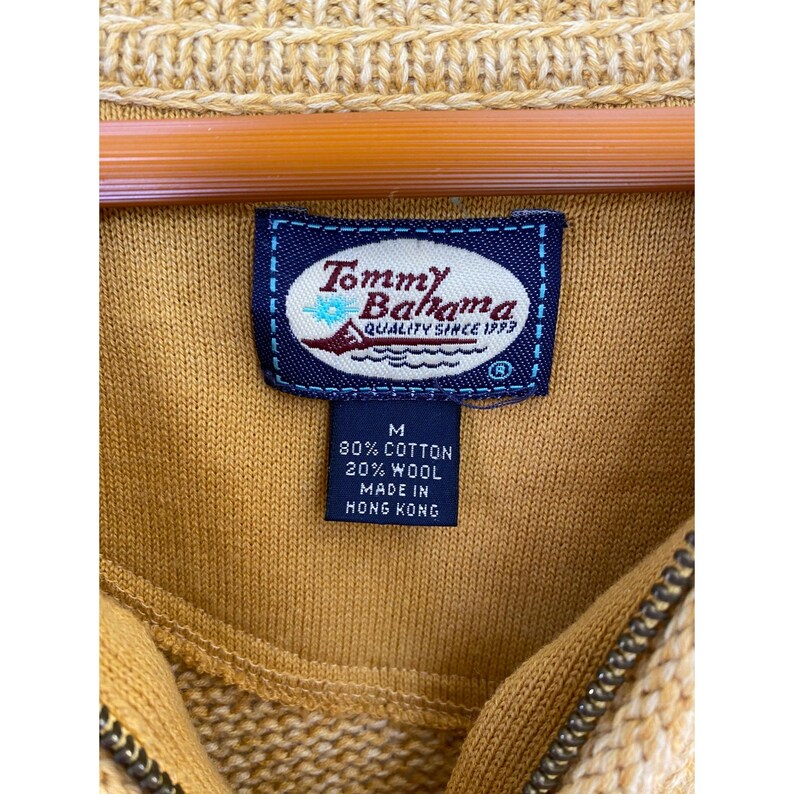 May include: Close-up of a mustard yellow sweater with a partial zipper and a label that reads "Tommy Bahama QUALITY SINCE 1993". The label also states the sweater is 80% cotton, 20% wool, and made in Hong Kong. The sweater is hanging on a wooden hanger.