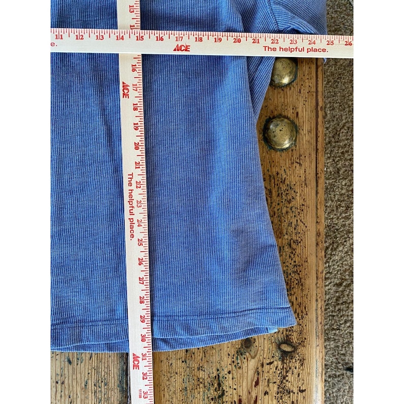 May include: A blue ribbed long-sleeve shirt featuring a horizontal and vertical Ace measuring tape. The horizontal tape shows a width of approximately 12 inches, while the vertical tape indicates a length of approximately 18 inches.