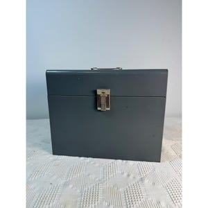 May include: A rectangular gray metal storage box featuring a smooth finish, a silver-toned metal latch centered on the front, and a small silver-toned handle attached to the top edge. The box has a clean, solid construction with visible metal seams.