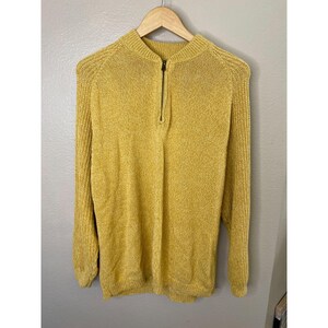 May include: Mustard yellow sweater with a half-zip closure. The long-sleeved sweater is hanging on a wooden hanger. The sweater has a ribbed texture on the shoulders and sleeves.