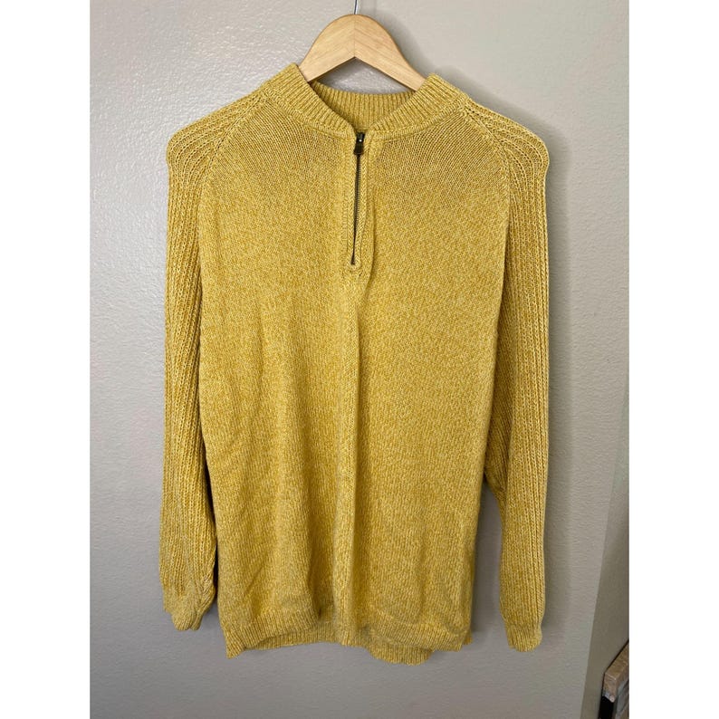 May include: Mustard yellow sweater with a half-zip closure. The long-sleeved sweater is hanging on a wooden hanger. The sweater has a ribbed texture on the shoulders and sleeves.