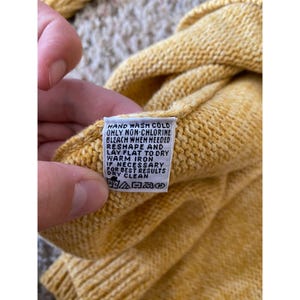 May include: Close-up of a yellow knit garment with a care label. The label provides washing instructions: hand wash cold, non-chlorine bleach when needed, reshape and lay flat to dry, warm iron if necessary, and dry clean for best results.