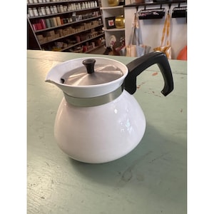 May include: White enamel coffee pot with a black handle and lid knob. The pot has a silver band around the middle and a spout for pouring. The handle is curved and the pot sits on a light green surface.