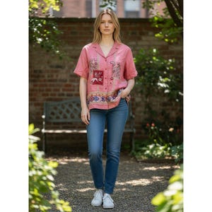 May include: A short-sleeved pink button-down shirt featuring a multi-colored paisley and floral pattern on the front, paired with blue denim jeans and white sneakers. The shirt has a collar and a button-up front closure with a straight hemline.