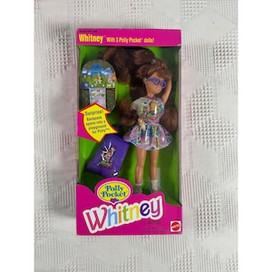 May include: A Whitney Polly Pocket doll set in original packaging. The doll has long brown hair, wears purple glasses and a patterned dress. Included is a purple backpack that opens into a playground. The box features the text Whitney with 3 Polly Pocket dolls and Polly Pocket logo.