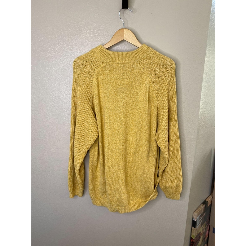 May include: Mustard yellow sweater with long sleeves and a ribbed texture. The sweater is hanging on a wooden hanger against a neutral wall. The sweater has a crew neck and a relaxed fit.