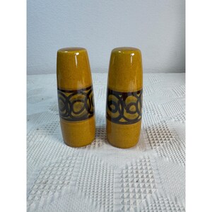 May include: A pair of cylindrical ceramic salt and pepper shakers featuring a glossy mustard yellow glaze. Each shaker is decorated with a wide, dark brown band around the center, which displays a repeating pattern of interlocking circular loops.
