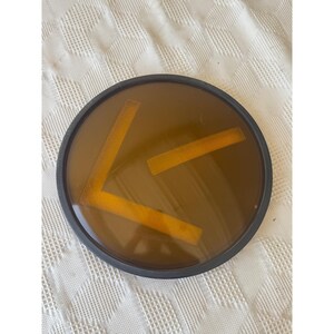 May include: A round, amber-colored lens with a black rim. Inside the lens are three rectangular, orange-colored shapes. The lens is set against a white, textured background.