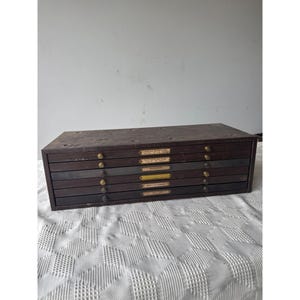 May include: A vintage wooden drawer cabinet with multiple drawers. The cabinet is dark brown with brass knobs and small labels above each drawer. The cabinet is rectangular and appears to be well-worn, suggesting age and use.