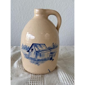 Puede incluir: Una jarra de cerámica beige con una sola asa y cuello estrecho. La parte frontal de la jarra presenta una ilustración pintada a mano en azul de una pequeña cabaña rodeada de árboles y una valla, con el número 99 pintado en azul abajo a la derecha.