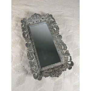 May include: A rectangular mirror featuring a grey, ornate frame with detailed relief carvings of leaves and flowers. The frame has a matte, weathered finish and surrounds a central, clear glass reflective surface with straight edges.
