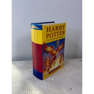 May include: A hardback book titled Harry Potter and the Order of the Phoenix by J.K. Rowling. The cover features a yellow background with a large orange and red phoenix rising from flames. The spine is yellow, red, and blue.
