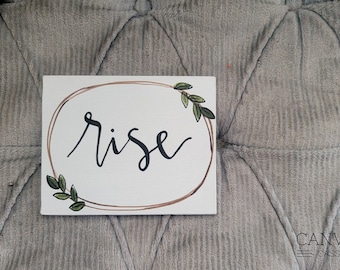May include: A white canvas sign with the word "rise" written in black calligraphy. The word is surrounded by a brown circle with green leaves on either side.