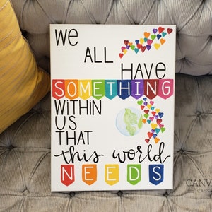 May include: A white canvas with the text "We all have something within us that this world needs" in black lettering. The text is surrounded by colorful hearts and a rainbow banner. The world is depicted in the center of the canvas.