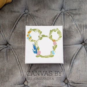 May include: A white canvas with a green floral Mickey Mouse silhouette. The flowers are yellow, blue, and pink.