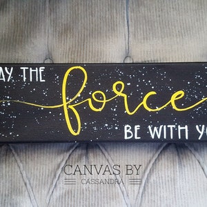 May include: A black canvas with white splatter paint and the words "May the force be with you" written in yellow paint. The canvas is signed "Canvas by Cassandra".