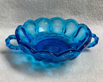 May include: A small, blue glass bowl with two handles. The bowl has a scalloped edge and a textured surface.