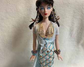 May include: A porcelain doll with black hair and blue eyes, wearing a white and turquoise beaded gown with gold accents. The doll is standing on a black base with the word "Gene" written on it.