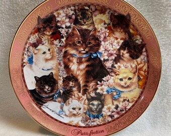 May include: A pink ceramic plate with a gold rim features a colorful illustration of multiple cats with blue ribbons. The plate has a gold inscription that reads "Purr-fection".