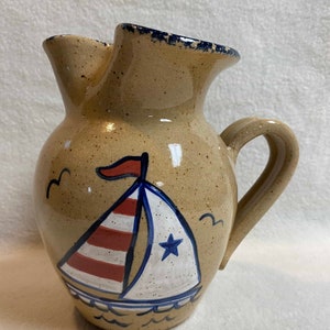 May include: Tan ceramic pitcher with a sailboat design. The sailboat has a white sail with red stripes and a blue star. The pitcher has a handle and a spout with a dark blue rim. Two birds are painted on the side.