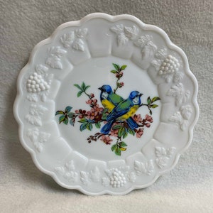 May include: White milk glass plate with a raised, embossed design of leaves and grapes around the edge. The center features a painted design of two blue birds perched on a branch with pink flowers.
