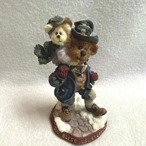 May include: A figurine of two teddy bears, one carrying the other on its back. The bears are dressed in winter attire, including hats, scarves, and coats. The base is inscribed with the words "GOD BLESS US. EVERYONE."