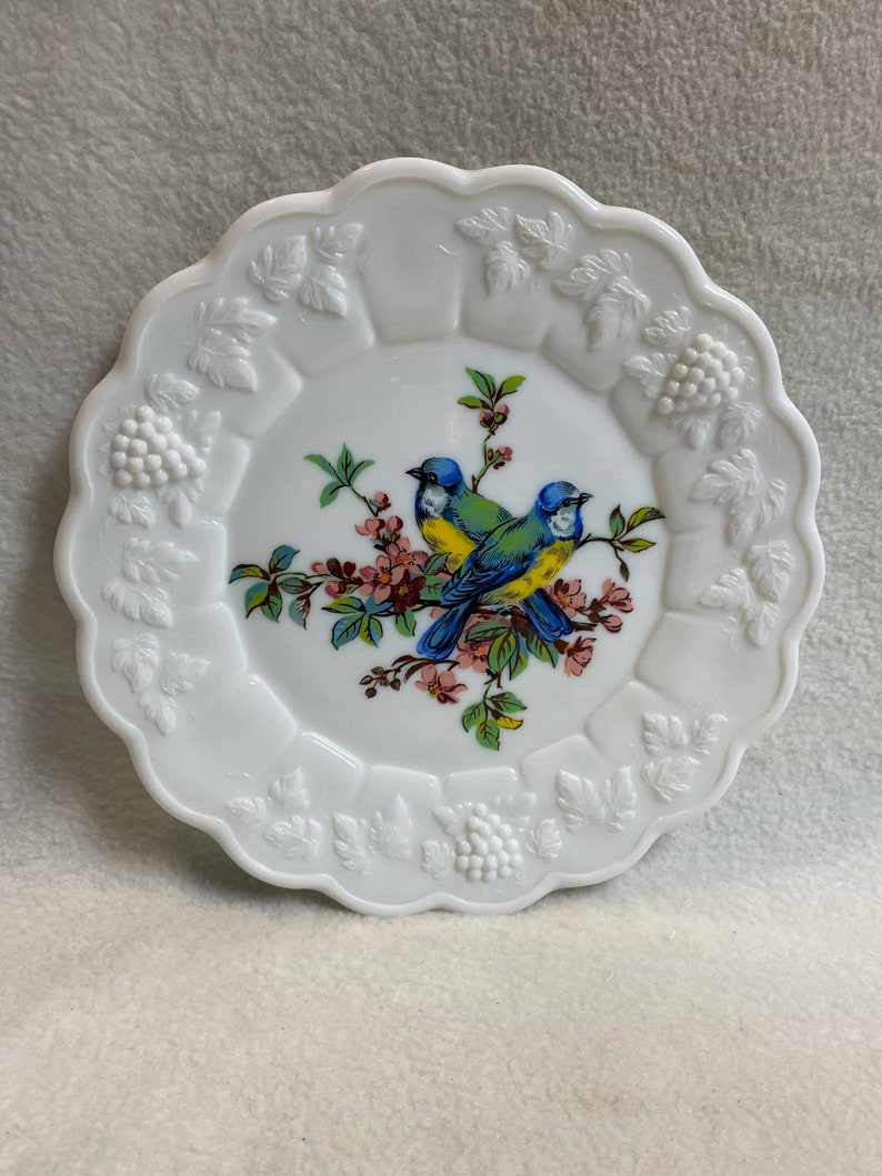 May include: White milk glass plate with a raised, embossed design of leaves and grapes around the edge. The center features a painted design of two blue birds perched on a branch with pink flowers.