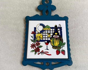 May include: Blue ceramic hot pad with a white tile featuring a red wine glass, a bottle of wine, cheese, and strawberries. The tile is surrounded by a blue frame with a decorative design.