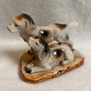 May include: A ceramic figurine of two brown and white dogs. The larger dog is standing on a brown base, while the smaller dog is lying down.