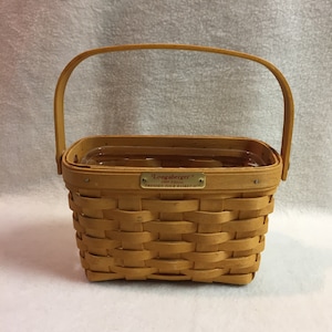 May include: A Longaberger Dresden Tour Basket II in a natural woven wicker with a clear plastic liner and a gold tag that reads "Longaberger 1999 Edition Dresden Tour Basket II"