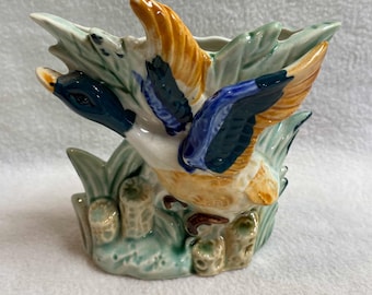 May include: A ceramic vase shaped like a duck flying over green reeds. The duck is painted in shades of blue, yellow, and orange. The vase is a vintage piece.