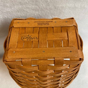 Może przedstawiać: Ręcznie tkany kosz Longaberger Baskets z brązowym wykończeniem z drewna. Kosz ma etykietę z napisem "Longaberger Baskets Handwoven Dresden Ohio USA". Kosz ma ręcznie napisany numer "65993" na spodzie.
