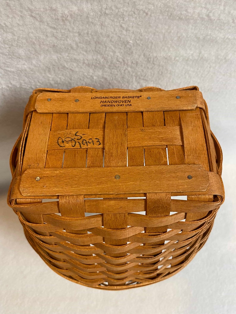 Może przedstawiać: Ręcznie tkany kosz Longaberger Baskets z brązowym wykończeniem z drewna. Kosz ma etykietę z napisem "Longaberger Baskets Handwoven Dresden Ohio USA". Kosz ma ręcznie napisany numer "65993" na spodzie.