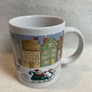 May include: White ceramic mug with a Christmas scene featuring a row of houses with smoke coming from the chimneys and Santa Claus riding in a sleigh pulled by reindeer.