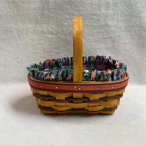 May include: A woven basket with a single upright handle, featuring a light tan, dark green, and deep red splint construction. The rim is accented with a dark red band and small silver-toned studs. A ruffled fabric liner with a floral pattern lines the interior.