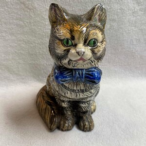 May include: A ceramic figurine of a cat wearing a blue bow tie. The cat has green eyes and a brown, white, and black striped pattern.