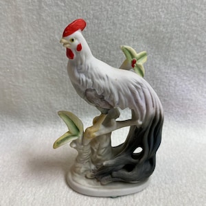 Pode incluir: Uma estatueta de cerâmica de um galo empoleirado num galho. O galo tem penas brancas com detalhes cinzentos, crista e barbela vermelhas, e é rodeado por detalhes florais verdes e vermelhos. A base é de cor cinza claro.