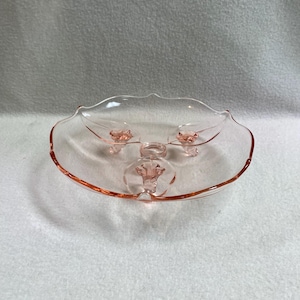 May include: A pink glass serving dish with a scalloped edge and three decorative feet. The dish has a shallow bowl shape and is made of transparent glass. The color is a soft, light pink, and the dish appears to be vintage.