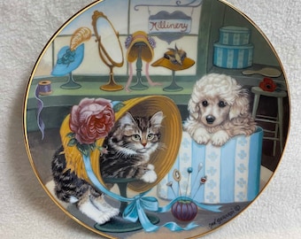 May include: A decorative ceramic plate featuring a whimsical illustration of a cat and a dog wearing hats in a millinery shop. The plate has a gold rim and the artist's signature "gaé guérin" in the bottom right corner.