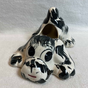 May include: A black and white ceramic dog figurine with a hole in the back. The dog is lying on its belly with its head turned to the side. The dog has large black eyes and a pink nose.