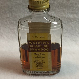 Pode incluir: Uma pequena garrafa de vidro transparente com uma tampa amarela e um rótulo amarelo. O rótulo diz "WATKINS COCONUT OIL SHAMPOO" e inclui o texto "Made from pure Coconut Oil" e "The J. R. Watkins Co.". A garrafa contém um líquido castanho.