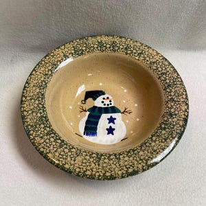 May include: A round ceramic bowl with a snowman design. The bowl has a tan interior with a hand-painted snowman wearing a black hat and a blue and green scarf. The rim is decorated with a speckled green pattern.