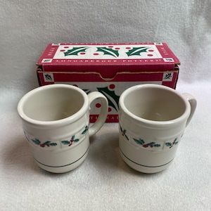 Op de afbeelding: Twee crèmekleurige keramische mokken met een patroon van groene hulstbladeren en rode bessen in het midden. Achter de mokken staat een rood-witte kartonnen doos met het opschrift Traditional Holly en Longaberger Pottery met hulstmotieven.