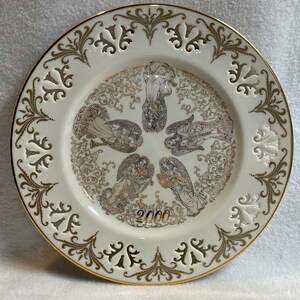 Op de afbeelding: Een wit porseleinen bord met een gouden rand en ingewikkelde gouden ontwerpen. Het midden van het bord toont een afbeelding van vijf engelen met gouden vleugels en een gouden inscriptie met de tekst "2000".