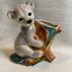 Pode incluir: Um vaso de cerâmica em forma de filhote de urso cinza sentado em um tronco de árvore marrom. O vaso tem um esmalte verde no interior e um esmalte marrom no exterior.