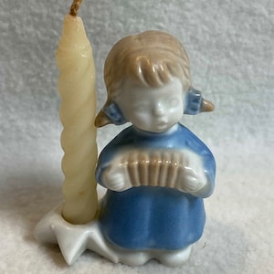 May include: A white ceramic candle holder featuring a girl figurine playing an accordion. The girl is wearing a blue dress and has brown hair. The candle holder has a white base and a white candle.
