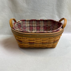 May include: A rectangular woven basket with a plaid fabric lining. The basket is made of light brown wood and has two curved leather handles. The plaid lining features red, white, and green colours.