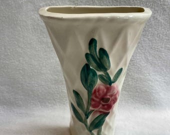 May include: Cream-colored ceramic vase with a textured diamond pattern. A painted floral design features a pink flower and green leaves. The vase has a flared shape and a rectangular opening, suitable for displaying flowers.