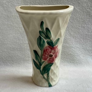 May include: A white ceramic vase with a diamond pattern and a pink flower with green leaves painted on the side. The vase has a curved top and a wide base.
