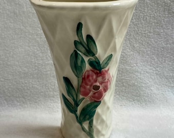 May include: A white ceramic vase with a diamond pattern and a pink flower with green leaves painted on the side. The vase has a curved top and a wide base.